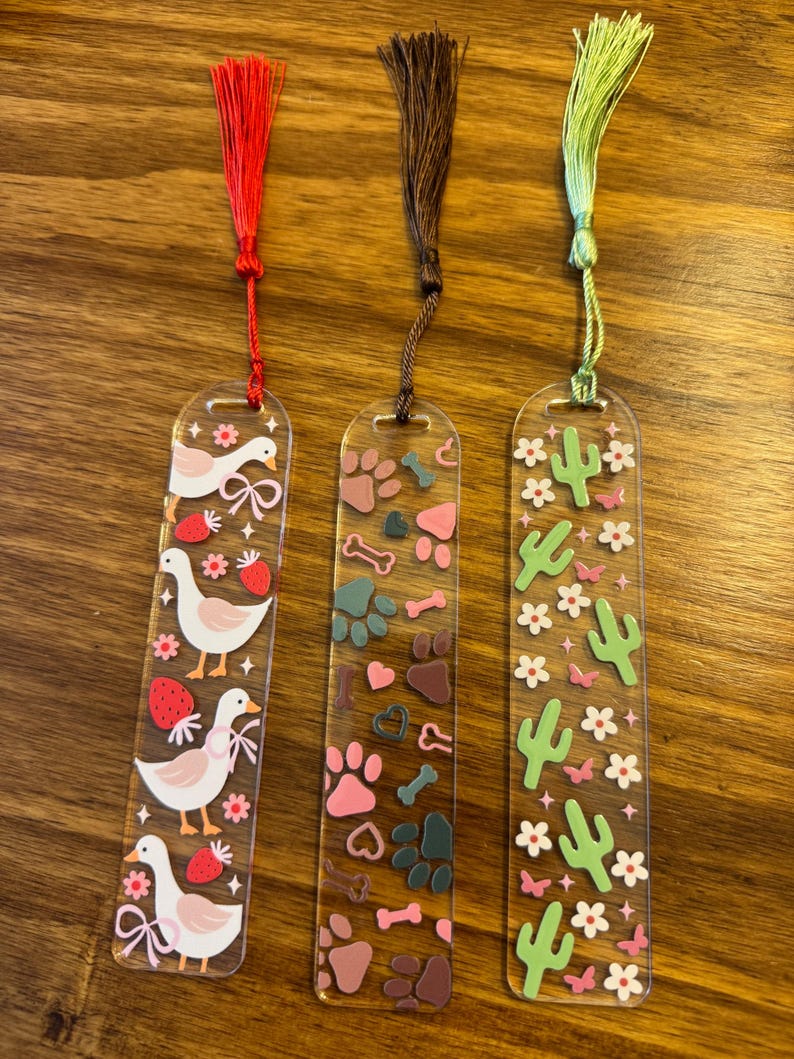 K&ouml;nnte beinhalten: Drei transparente Acryl-Lesezeichen mit farbenfrohen Designs und Quasten. Eines zeigt G&auml;nse, Erdbeeren und Schleifen; ein anderes Pfotenabdr&uuml;cke, Herzen und Knochen; das letzte Kakteen und Blumen. Jedes Lesezeichen hat eine andersfarbige Quaste.