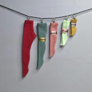 May include: Five miniature fabric stockings in varying lengths and colours, including red, sage green, dusty pink, light green, and mustard yellow, are suspended from a black cord by silver metal clips. Each stocking features a small label with the text handmade.