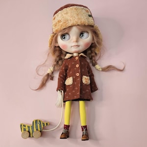 May include: A doll with light brown hair in braids, wearing a brown patterned coat, yellow tights, brown lace-up boots, and a fuzzy brown hat. The doll holds a string attached to a small, yellow and black striped pull-toy duck on wheels.