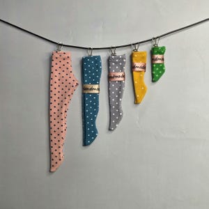 May include: Five fabric leg-shaped ornaments hanging from a black wire by metal clips. The ornaments are pink, teal, grey, yellow, and green, all featuring white or black polka dots. Each piece has a small ribbon label printed with the word handmade.