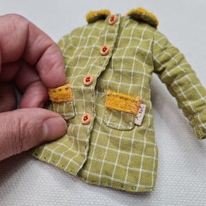 May include: A miniature olive green jacket with a white grid pattern, featuring a mustard yellow collar and pocket flaps. The garment has small red buttons down the front and a small white fabric tag on the right pocket with the letters a.w. printed.