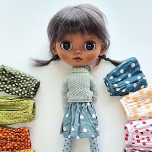 May include: A doll with dark grey hair in pigtails, large dark eyes, and a light brown complexion. The doll wears a light green knit jumper, a teal skirt with white polka dots, and blue polka-dot tights, surrounded by various fabric sleeves.