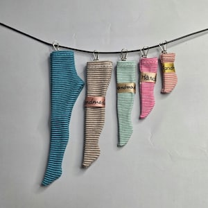 May include: Five pairs of miniature striped stockings in varying sizes and colours, including blue, tan, mint green, pink, and light pink, are suspended from a black wire by silver metal clips. Each stocking features a small tag labelled handmade.