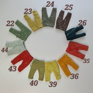 May include: A collection of eleven pairs of small trousers arranged in a circle, each featuring a drawstring waist, side pockets, and a small label. The trousers are numbered 22, 23, 24, 25, 26, 35, 36, 39, 40, 43, and 44 in various colours including green, yellow, brown, and red.