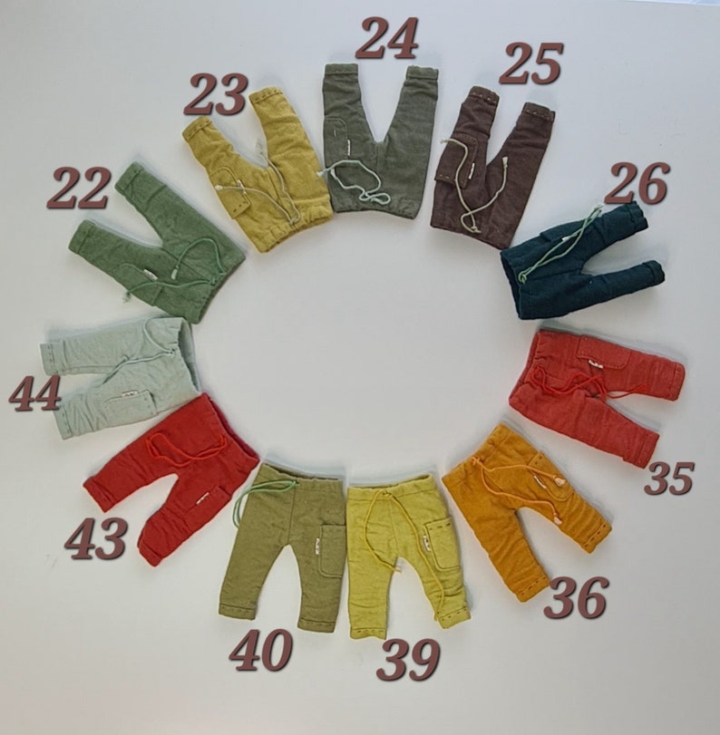 May include: A collection of eleven pairs of small trousers arranged in a circle, each featuring a drawstring waist, side pockets, and a small label. The trousers are numbered 22, 23, 24, 25, 26, 35, 36, 39, 40, 43, and 44 in various colours including green, yellow, brown, and red.