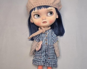 Puede incluir: Una muñeca con cabello azul oscuro que lleva una boina de punto beige, un mono de manga corta a rayas azules y blancas sobre una camiseta de manga larga con rayas horizontales finas, un pequeño bolso cruzado rosa, leggings naranjas y calcetines color canela.