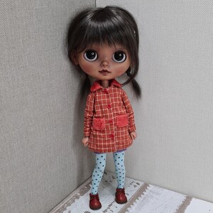 May include: A doll with dark brown hair and large eyes wearing an orange and white plaid coat with red buttons and two fuzzy red pockets. The doll is dressed in light blue leggings with a dark blue polka dot pattern and small brown shoes.