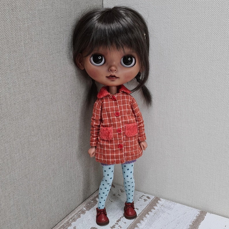 May include: A doll with dark brown hair and large eyes wearing an orange and white plaid coat with red buttons and two fuzzy red pockets. The doll is dressed in light blue leggings with a dark blue polka dot pattern and small brown shoes.