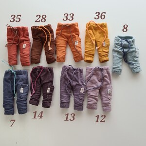 May include: A collection of nine small doll trousers in various colours including rust, brown, orange, yellow, light blue, dark grey, dark brown, purple, and lavender. Each pair features a drawstring waist, a side pocket, and a small white label.