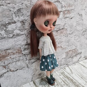 May include: A doll with long brown hair wearing a white blouse with a lace trim and a green and white polka dot skirt. The doll is wearing black shoes and has green eyes.