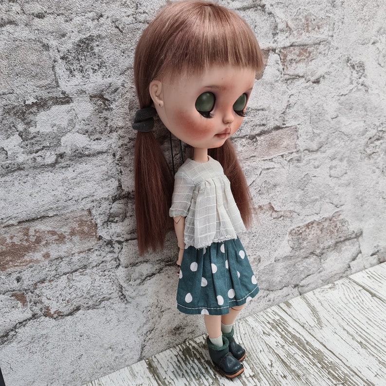 May include: A doll with long brown hair wearing a white blouse with a lace trim and a green and white polka dot skirt. The doll is wearing black shoes and has green eyes.