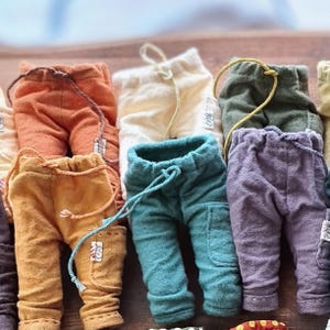 May include: A collection of small, textured fabric trousers in various colours including orange, cream, olive green, mustard yellow, teal, and purple. Each pair features an elastic waistband with a drawstring, side pockets, and a small white brand label.