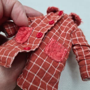 May include: A miniature long-sleeved jacket in a rust-orange fabric with a white grid pattern. The jacket features a small collar, four bright red buttons down the front, and two textured, fuzzy red patch pockets on the lower front panels.