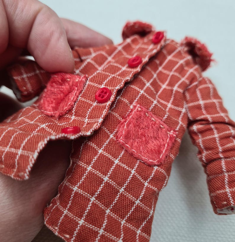 May include: A miniature long-sleeved jacket in a rust-orange fabric with a white grid pattern. The jacket features a small collar, four bright red buttons down the front, and two textured, fuzzy red patch pockets on the lower front panels.