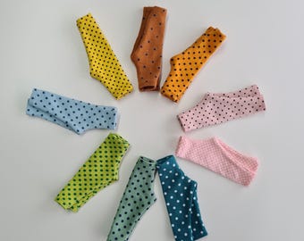 Peut inclure: Un ensemble de neuf leggings miniatures disposés en cercle. Chaque paire présente une couleur unie distincte, notamment jaune, orange, bleu clair, vert, sarcelle et rose, toutes ornées d'un motif répétitif de petits points foncés.