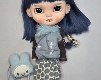Puede incluir: Una muñeca con cabello azul oscuro, ojos negros grandes y mejillas sonrosadas. Viste una sudadera gris con estampado de piñas blancas, una camiseta marrón de manga larga y una falda gris con lunares blancos grandes. Incluye un pequeño bolso de peluche azul.