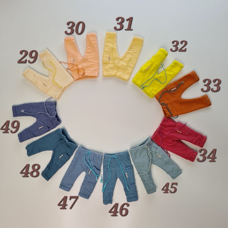 May include: A circular arrangement of eleven pairs of small trousers in various colours, each labelled with a number from 29 to 49. The trousers feature elastic waistbands with drawstrings, side pockets, and stitched detailing on the cuffs and hems.