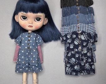 May include: A doll with dark blue hair wearing a blue polka-dot denim romper over a pink and white striped long-sleeve shirt, beige socks, and brown shoes, displayed next to a stack of five additional denim rompers in various patterns.