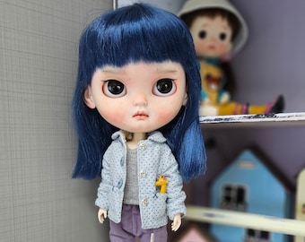 May include: A blue-haired doll with a light blue jacket and grey shirt. The doll has a serious expression and is wearing purple pants.