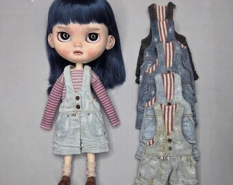 May include: A doll with dark blue hair, wearing a pink and purple striped long-sleeve shirt, light blue distressed denim overalls, beige socks, and brown shoes, displayed next to a stack of four additional pairs of denim overalls in various shades.