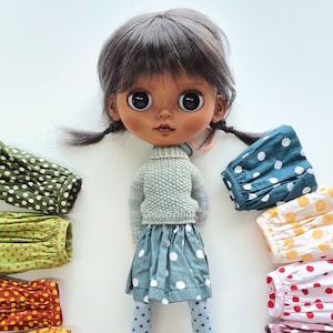 May include: A doll with dark brown hair and brown eyes wearing a light green sweater and a blue and white polka dot skirt. The doll is surrounded by a variety of colorful polka dot skirts in different colors, including green, yellow, red, and orange.