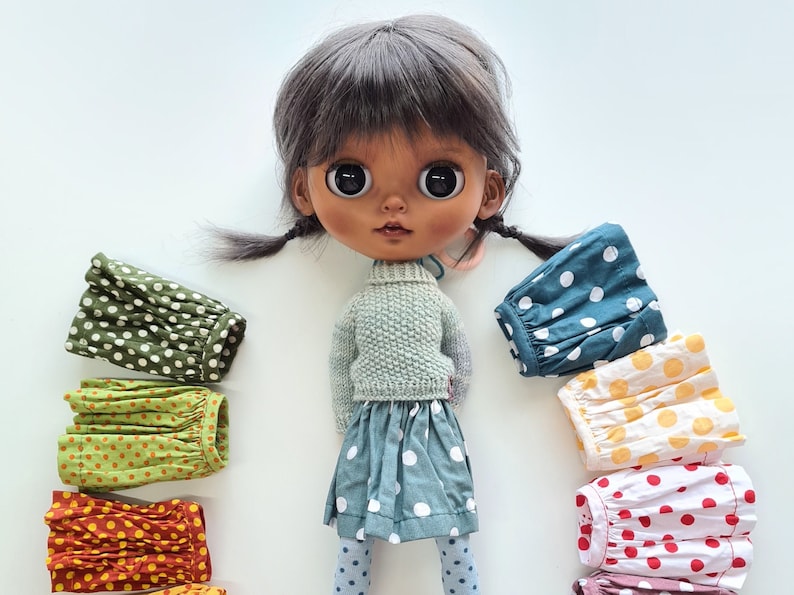 May include: A doll with dark brown hair and brown eyes wearing a light green sweater and a blue and white polka dot skirt. The doll is surrounded by a variety of colorful polka dot skirts in different colors, including green, yellow, red, and orange.