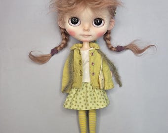 Puede incluir: Una muñeca con cabello castaño claro peinado en dos trenzas, ojos grandes y oscuros, y mejillas sonrosadas. La muñeca viste una chaqueta verde claro sobre una camisa blanca, falda verde de lunares, medias a rayas amarillas y grises y zapatos verdes.