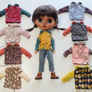 May include: A doll with dark hair in pigtails wearing a yellow polka-dot hoodie, teal sleeves, and grey trousers, surrounded by eight miniature hoodies in various colours and patterns including floral, polka-dot, and fruit prints.