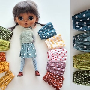 May include: A doll wearing a blue polka dot skirt and tights, surrounded by a rainbow of colorful polka dot skirts. The skirts are made of fabric with a pleated design.
