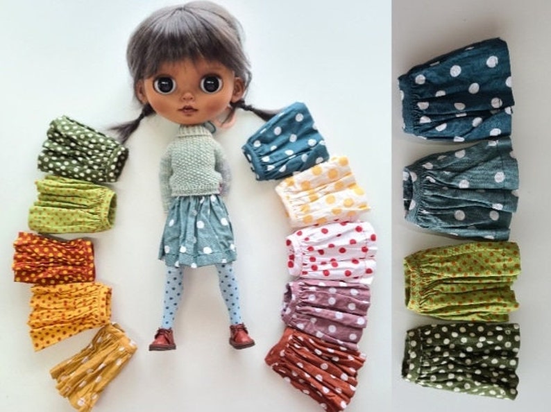 May include: A doll wearing a blue polka dot skirt and tights, surrounded by a rainbow of colorful polka dot skirts. The skirts are made of fabric with a pleated design.