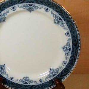 May include: White ceramic plate with a blue and gold decorative border. The plate features an intricate blue pattern around the rim and a gold trim. The plate is displayed on a wooden stand.