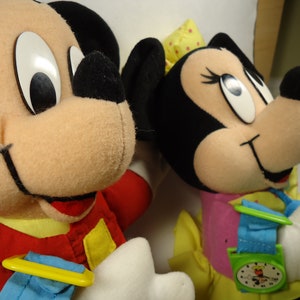 May include: Two plush toys, Mickey Mouse and Minnie Mouse, are dressed in red and pink outfits. Mickey Mouse is wearing a red jacket with yellow accents and a blue and yellow watch. Minnie Mouse is wearing a pink dress with yellow accents and a blue and green watch.