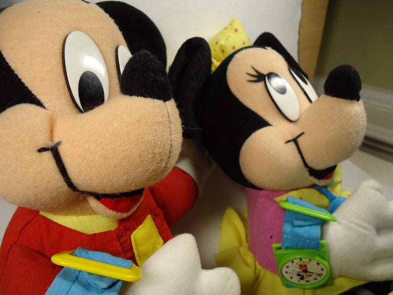 May include: Two plush toys, Mickey Mouse and Minnie Mouse, are dressed in red and pink outfits. Mickey Mouse is wearing a red jacket with yellow accents and a blue and yellow watch. Minnie Mouse is wearing a pink dress with yellow accents and a blue and green watch.