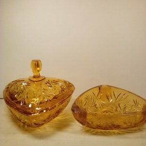 May include: Two amber-colored glass heart-shaped dishes. One is a covered candy dish with a lid and a small handle. The other is an open dish. Both have a leaf and swirl pattern. The dishes are on a white surface.
