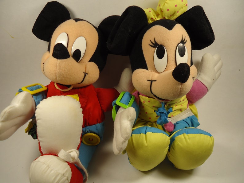 May include: A pair of plush Mickey Mouse and Minnie Mouse toys. Mickey is wearing a red, blue, and yellow outfit. Minnie is wearing a yellow, blue, and pink outfit with white polka dots.