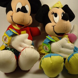 May include: Two plush toys, Mickey Mouse and Minnie Mouse, are wearing colorful outfits and have watches on their wrists. Mickey Mouse is wearing a red, yellow, and blue outfit with a black and white checkered pattern. Minnie Mouse is wearing a yellow, blue, and pink outfit with white polka dots.