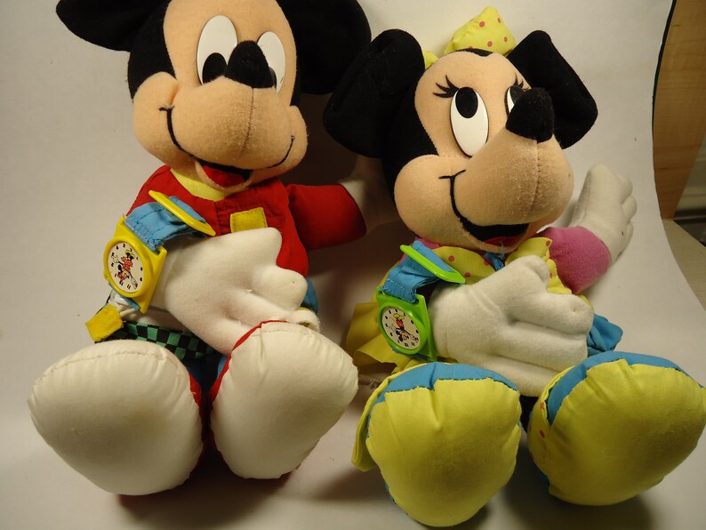 May include: Two plush toys, Mickey Mouse and Minnie Mouse, are wearing colorful outfits and have watches on their wrists. Mickey Mouse is wearing a red, yellow, and blue outfit with a black and white checkered pattern. Minnie Mouse is wearing a yellow, blue, and pink outfit with white polka dots.