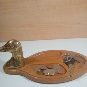 May include: A wooden duck-shaped trinket tray with a brass duck head on the left side. The tray has three compartments, one for keys, one for coins, and one for other small items. The tray is made of light brown wood and has a natural finish.