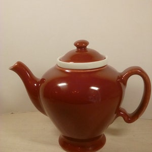 May include: A brown ceramic teapot with a white rim on the lid. The teapot has a rounded body and a spout on the side.