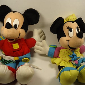 May include: A pair of plush Mickey Mouse and Minnie Mouse toys. Mickey is wearing a red, blue, and yellow racing suit. Minnie is wearing a pink, yellow, and blue dress with white polka dots.