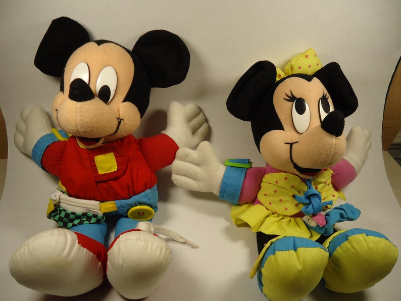 May include: A pair of plush Mickey Mouse and Minnie Mouse toys. Mickey is wearing a red, blue, and yellow racing suit. Minnie is wearing a pink, yellow, and blue dress with white polka dots.