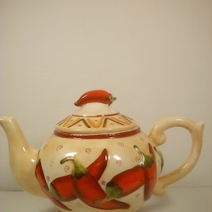 May include: A white ceramic teapot with a red chili pepper lid and red chili pepper designs on the body. The teapot has a brown, orange, and white pattern around the base.
