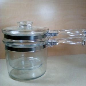 May include: A clear glass double boiler with a glass lid and two clear handles. The double boiler has a stainless steel band around the middle and is designed for cooking delicate foods. The pot is sitting on a light-colored surface.