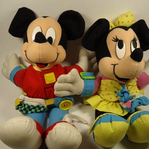 May include: A pair of plush Mickey Mouse and Minnie Mouse toys. Mickey is wearing a red jacket, blue jeans, and a checkered belt. Minnie is wearing a yellow dress with pink polka dots and a blue bow.