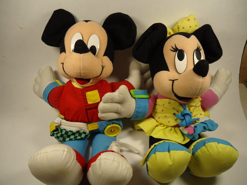 May include: A pair of plush Mickey Mouse and Minnie Mouse toys. Mickey is wearing a red jacket, blue jeans, and a checkered belt. Minnie is wearing a yellow dress with pink polka dots and a blue bow.