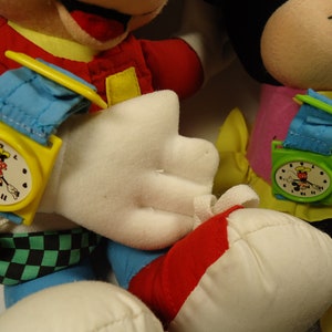 May include: Two plush Mickey Mouse toys wearing colorful watches with Mickey Mouse faces on the watch faces. The Mickey Mouse on the left is wearing a yellow watch with a blue strap and the Mickey Mouse on the right is wearing a green watch with a blue strap.