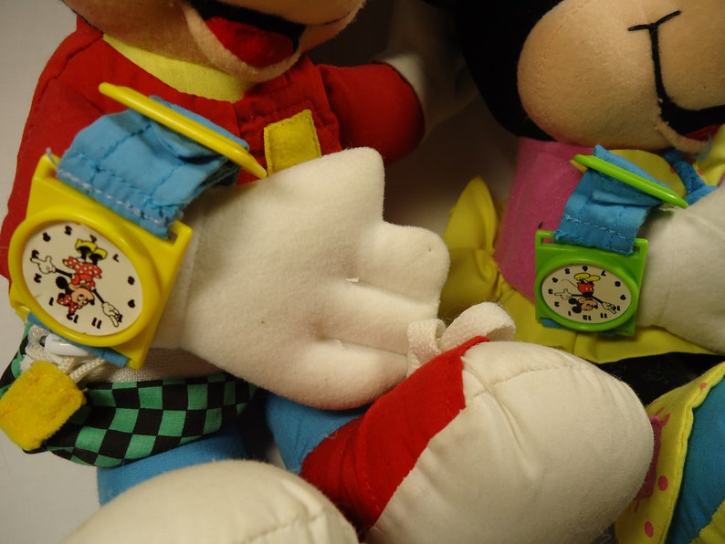 May include: Two plush Mickey Mouse toys wearing colorful watches with Mickey Mouse faces on the watch faces. The Mickey Mouse on the left is wearing a yellow watch with a blue strap and the Mickey Mouse on the right is wearing a green watch with a blue strap.