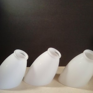 May include: Three white glass lamp shades with a rounded, conical shape and a flared opening. The shades are arranged in a row, slightly angled, against a dark background. The shades are opaque and appear to be made of a smooth glass material.