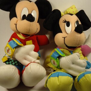 May include: A pair of plush Mickey Mouse and Minnie Mouse toys. Mickey is wearing a red, yellow, and blue outfit with a watch on his wrist. Minnie is wearing a yellow, blue, and pink outfit with a watch on her wrist.