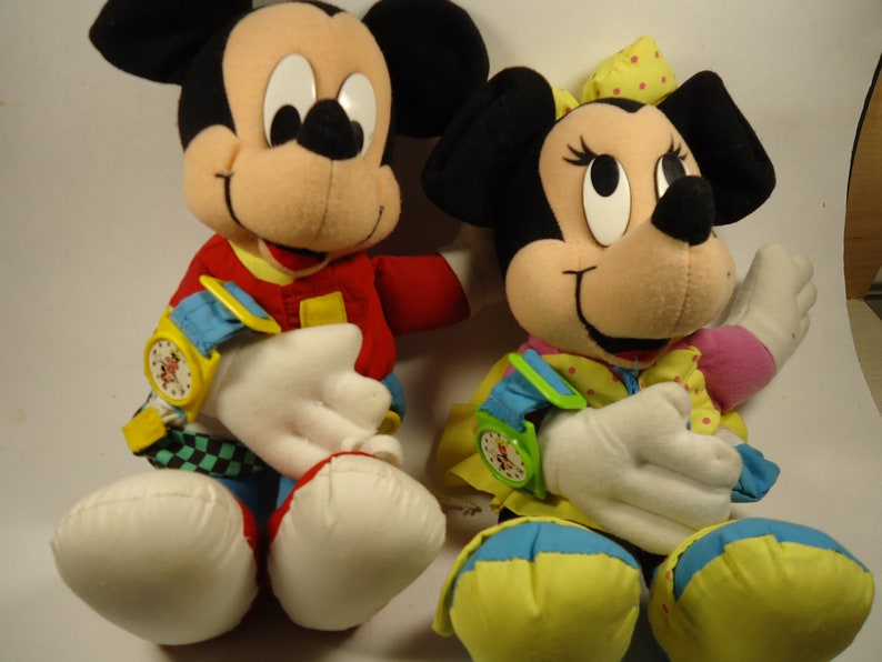 May include: A pair of plush Mickey Mouse and Minnie Mouse toys. Mickey is wearing a red, yellow, and blue outfit with a watch on his wrist. Minnie is wearing a yellow, blue, and pink outfit with a watch on her wrist.
